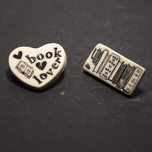 May include: Two off-white pins. One is heart-shaped with the words "book lover" and a small book illustration. The other is rectangular with a stack of books illustrated in black. Both pins have a glossy finish.