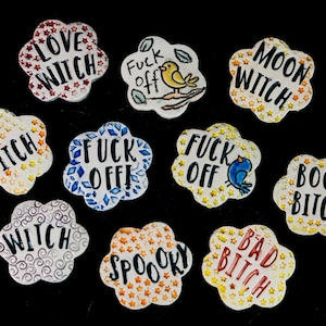 May include: Ten ceramic flower-shaped ornaments with various sayings, including "Love Witch", "Moon Witch", "Boo Bitch", "Bad Bitch", "Spooky", "Witch", and "Fuck Off".