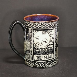 May include: A ceramic mug with a black handle and rim. The mug features a black and white design with the text "THE SUN" and a smiling sun illustration. The interior rim is a dark purple color.