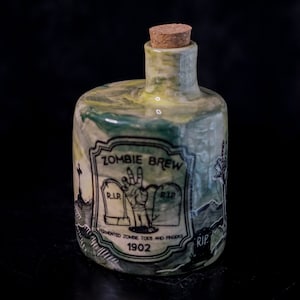 Puede incluir: Una pequeña botella de cerámica verde y amarilla con un tapón de corcho. La botella tiene una etiqueta que dice "Zombie Brew Fermented Zombie Toes and Fingers 1902" con un dibujo de una mano de zombie y una lápida.