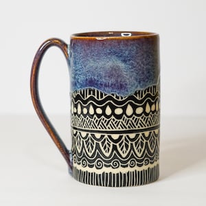 May include: A ceramic mug featuring a glossy purple and blue gradient glaze on the upper portion and a black lower section decorated with intricate white hand-carved patterns including waves, leaves, and geometric lines with a curved handle.
