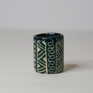 May include: A cylindrical ceramic cup featuring a deep teal glaze with contrasting cream-colored geometric patterns. The exterior surface is decorated with vertical bands of repeating wavy lines, chevrons, and diagonal hatch marks.