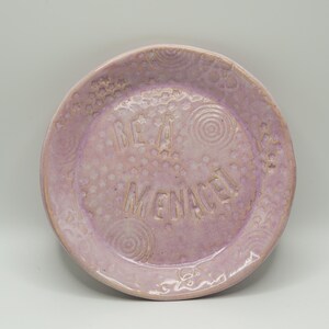 May include: A handmade, lavender-colored ceramic plate with the words "BE A MENACE" inscribed in the center. The plate features a textured surface with dotted and spiral patterns around the rim.