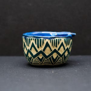 May include: A small ceramic bowl with a blue rim and a geometric pattern of green and white triangles and lines.