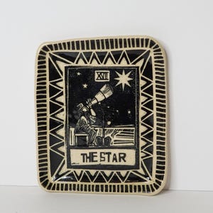 May include: A rectangular ceramic tray featuring a black and cream color scheme. The center depicts a figure looking through a telescope under a large star with the Roman numeral XVII and the text THE STAR. A geometric border frames the design.