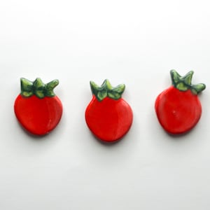 May include: Three ceramic strawberry-shaped magnets. Each magnet has a glossy red body and a green leafy top. The magnets are arranged in a horizontal line against a white background.