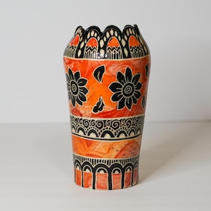 May include: A ceramic vase featuring a vibrant orange and red marbled background with black hand-painted floral motifs and intricate geometric patterns. The rim has a scalloped edge, and the body displays horizontal bands of decorative arches.
