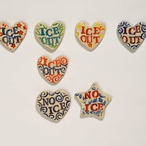 May include: A collection of ceramic heart and star-shaped magnets. The magnets are decorated with the words "ICE OUT" and "NO ICE" in various colors and patterns, including red, blue, green, and yellow. The magnets are likely for use on a refrigerator or other magnetic surface.
