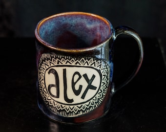 May include: A black ceramic mug with a white design. The design features a decorative border and the name "Alex" in a stylized font.