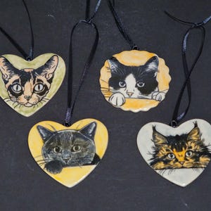 May include: Four ceramic cat ornaments with black ribbon hangers. Each ornament features a different cat portrait. Two are heart-shaped, one is round with a scalloped edge, and all have a yellow or light green background.