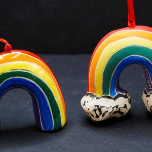 May include: Two rainbow-colored ceramic ornaments shaped like rainbows with clouds at the ends. The rainbows are painted in red, orange, yellow, green, blue, indigo, and violet. The clouds are white with black details. One ornament has a red ribbon attached.