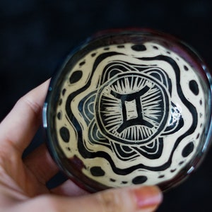 May include: A white ceramic bowl with a black and white mandala design featuring the Gemini zodiac symbol. The bowl has a dark brown rim.