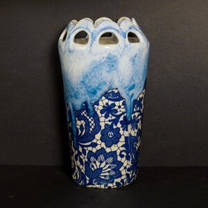 May include: A tall ceramic vase with a unique design. The top portion is white with blue watercolor-like drips, and the bottom features a blue and white floral lace pattern. The rim has a scalloped edge with evenly spaced openings.