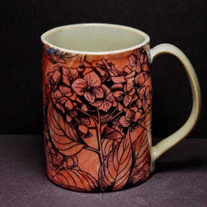 May include: A ceramic mug with a floral design. The mug features a peach-colored background with a black floral outline. The handle and interior are a light green color. The mug is a kitchen and dining item.