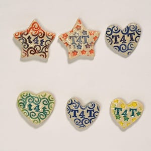 May include: Six ceramic decorative pieces in star and heart shapes. The stars are red and orange with a swirl design and the text "T4T". The hearts are blue, green, and yellow with the text "T4T" and a swirl design.