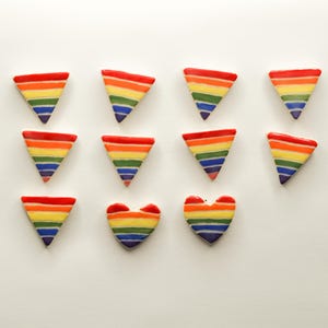 May include: Overhead shot of a collection of colorful, rainbow-striped ceramic shapes. The shapes include triangles and hearts, each with a red top and a gradient of yellow, orange, green, blue, and purple stripes. The shapes are arranged on a white surface.