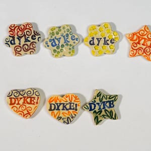 May include: Seven ceramic pins in various shapes including flowers, stars, and hearts. Each piece features colorful patterns and the word dyke or DYKE! in blue, red, or black lettering. The designs include swirls, dots, stars, and leaf motifs.