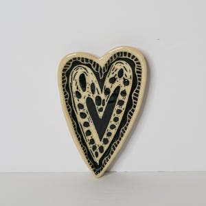 May include: A heart-shaped ceramic dish featuring a glossy cream-colored border and a central black interior. The surface is decorated with intricate black hand-painted patterns, including dots and lines that follow the heart's contour.