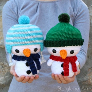 Puede incluir: Dos juguetes de muñeco de nieve de crochet, uno con un gorro a rayas azul y blanco y una bufanda azul marino, el otro con un gorro verde y una bufanda roja.