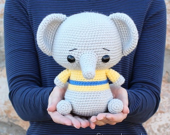 May include: A gray crocheted elephant wearing a yellow and blue striped shirt. The elephant has large black eyes and a small trunk.
