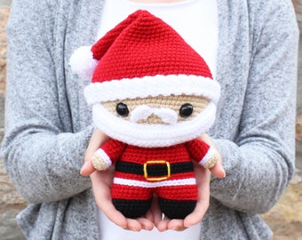 Puede incluir: Un muñeco de Santa Claus de crochet con un gorro rojo, barba blanca y botas negras. El muñeco lleva una bolsa roja con ribete blanco.