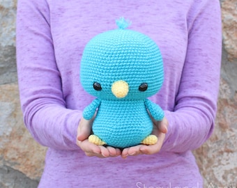 May include: A crocheted blue bird with yellow beak and feet. The bird has black eyes and a small tuft of blue yarn on its head.