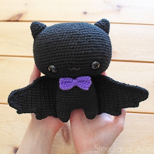 May include: A black crocheted bat with a purple bow tie. The bat has large eyes and a small, cute smile.