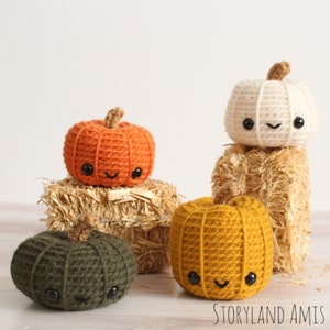 May include: Four crocheted pumpkins in different colors: orange, white, mustard yellow, and dark green. Each pumpkin has a cute, smiling face with black eyes and a stitched-on mouth. The pumpkins are sitting on a bed of straw.