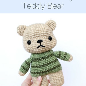 May include: A crocheted teddy bear wearing a green and brown striped sweater. The bear has black eyes and a black nose. The bear is being held by a hand. The text "Edmond the Cozy Teddy Bear" is at the top of the image. The text "www.storylandamis.com" is at the bottom of the image.