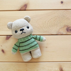 May include: A crocheted teddy bear wearing a green and brown striped sweater. The bear has black eyes and a black nose.