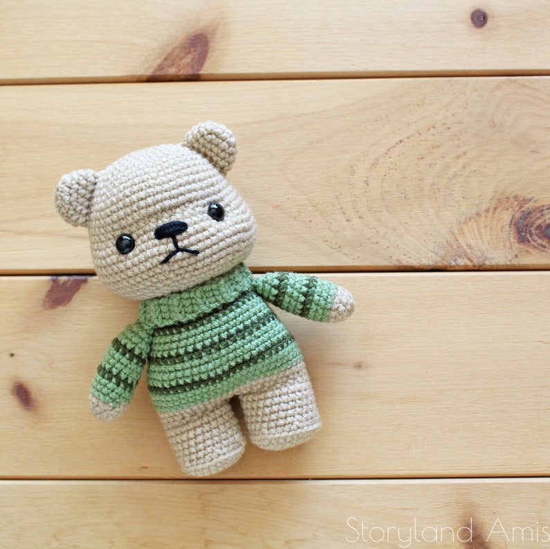May include: A crocheted teddy bear wearing a green and brown striped sweater. The bear has black eyes and a black nose.