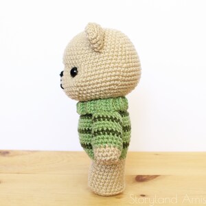 May include: A crocheted teddy bear wearing a green and brown striped sweater. The bear is standing on a wooden surface.