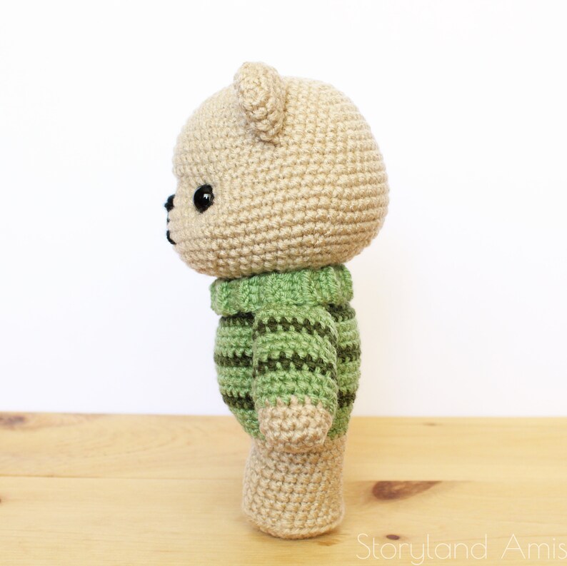 May include: A crocheted teddy bear wearing a green and brown striped sweater. The bear is standing on a wooden surface.