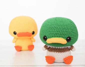 Puede incluir: Dos peluches de pato de crochet, uno amarillo y otro verde con detalles marrones. El pato amarillo tiene patas y pico naranja. El pato verde tiene ojos negros y pico naranja.