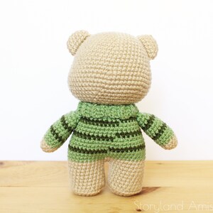 May include: A crocheted teddy bear wearing a green and brown striped sweater. The bear is standing on a wooden surface.