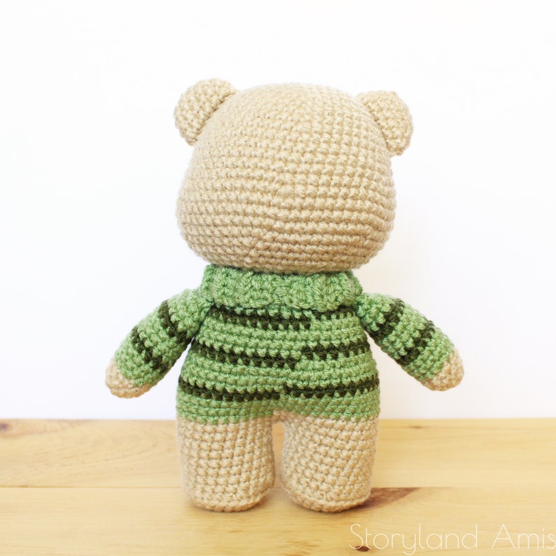May include: A crocheted teddy bear wearing a green and brown striped sweater. The bear is standing on a wooden surface.