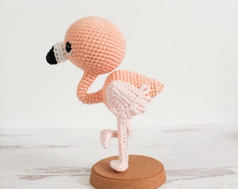 May include: A pink and white crocheted flamingo toy standing on a cork base. The flamingo has a black eye and a white beak.