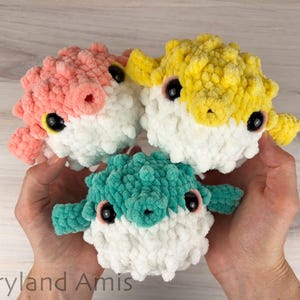 May include: Three plush pufferfish toys made of soft yarn, each featuring a white underbelly and a colored top in shades of pastel pink, bright yellow, and teal green. Each pufferfish has large black plastic eyes and a small circular mouth.