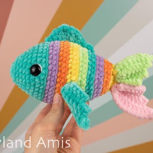 May include: A colorful, crocheted fish toy with a turquoise head, rainbow body stripes, and a pink and green tail. The fish has large black eyes and is held in a hand. The background is a striped pattern of various colors.