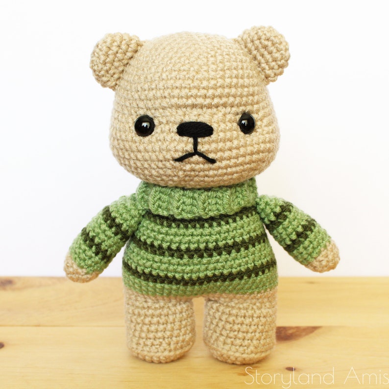 May include: A crocheted teddy bear wearing a green and brown striped sweater. The bear has black eyes and a black nose.