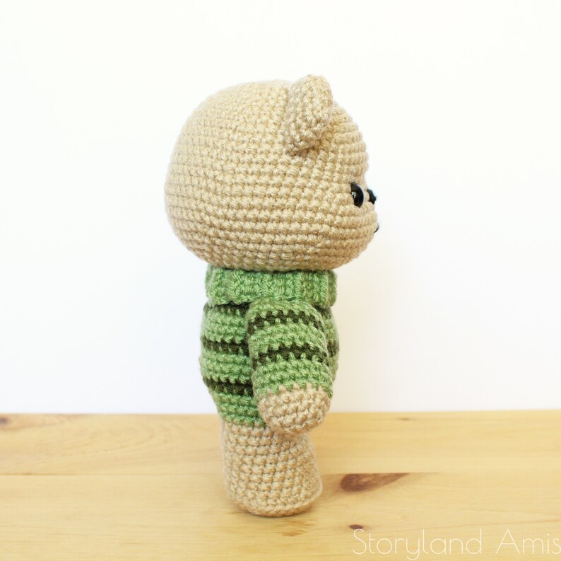 May include: A crocheted teddy bear wearing a green and brown striped sweater. The bear is standing on a wooden surface.