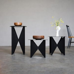 May include: Three black metal side tables with a geometric design, each topped with a wooden bowl. The tables are arranged in a row, with a chair and a red and orange abstract painting in the background.