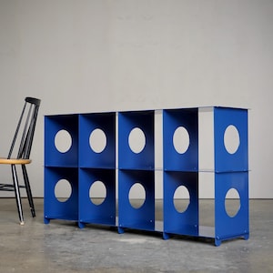 May include: A blue storage unit with eight cubbies, each featuring a large, oval cutout. A black chair with a wooden seat and back is positioned to the left of the unit. The unit is made of metal and has a modern design.