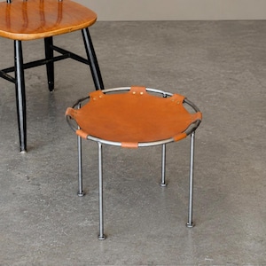 May include: A circular stool featuring a tan leather seat stretched over a silver-toned metal frame with four thin legs. The leather is secured to the circular frame with small metal rivets. A wooden chair with black legs is partially visible.