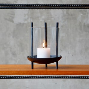 May include: A black metal candle holder with a clear glass cylinder and a white candle. The candle holder is on a wooden shelf.