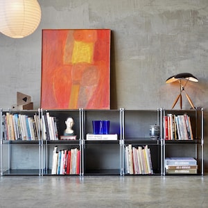 May include: A metal shelving unit with five shelves, filled with books and decorative items. The shelves are against a gray wall with a painting and a lamp on top.