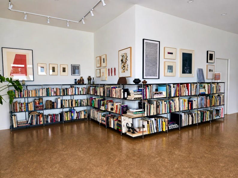May include: A well-lit room with a large collection of books displayed on black metal shelving units. Various framed artworks adorn the white walls. The floor is a warm, light brown color.