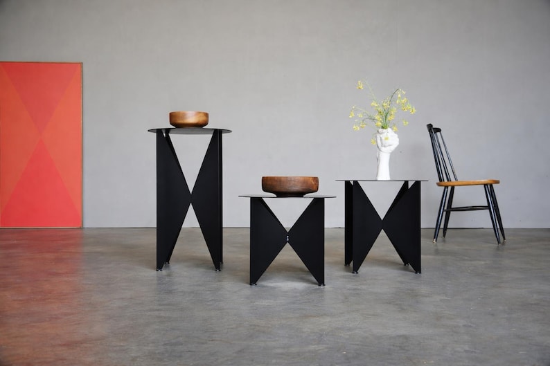 Puede incluir: Tres mesas auxiliares de metal negro con un dise&ntilde;o geom&eacute;trico, cada una coronada con un cuenco de madera. Las mesas est&aacute;n dispuestas en fila, con una silla y una pintura abstracta roja y naranja en el fondo.