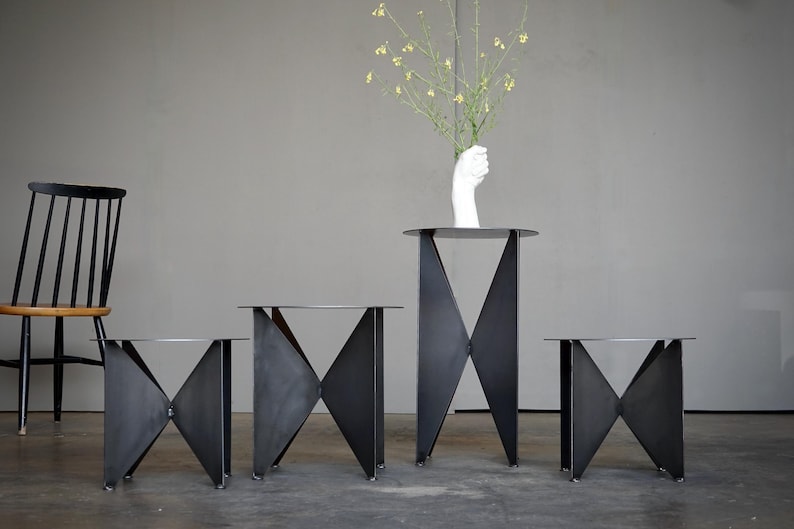 Puede incluir: Un conjunto de cuatro mesas auxiliares de metal negro con un dise&ntilde;o geom&eacute;trico &uacute;nico. Las mesas tienen una parte superior plana y circular y una base formada por piezas de metal que se cruzan. Una mesa m&aacute;s alta sostiene un jarr&oacute;n blanco en forma de mano con flores amarillas.