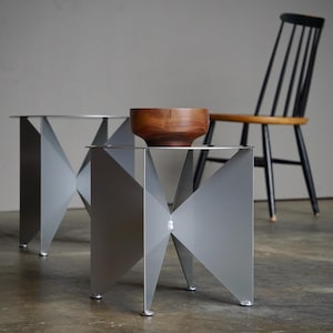 Puede incluir: Dos mesas auxiliares modernas y geom&eacute;tricas con acabado plateado. Cada mesa tiene un dise&ntilde;o angular &uacute;nico y una parte superior circular. Un cuenco de madera descansa sobre una mesa. Una silla negra con asiento de madera est&aacute; al fondo.